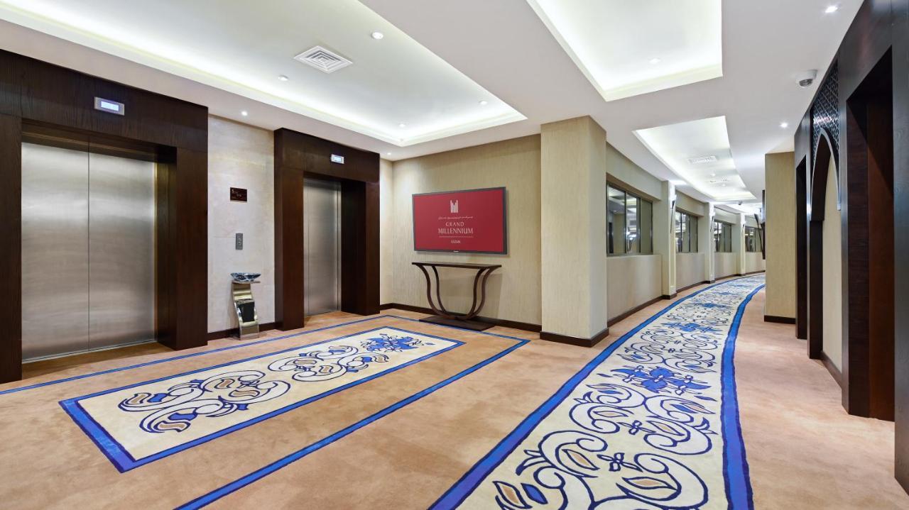 Hotel Grand Millennium Gizan à Jizan Extérieur photo The photo shows a hotel corridor featuring two elevator doors on the left side. To the right, there is a wall with a red sign, likely indicating the name or logo of the hotel. The floor is adorned with a decorative carpet that has a blue and beige de